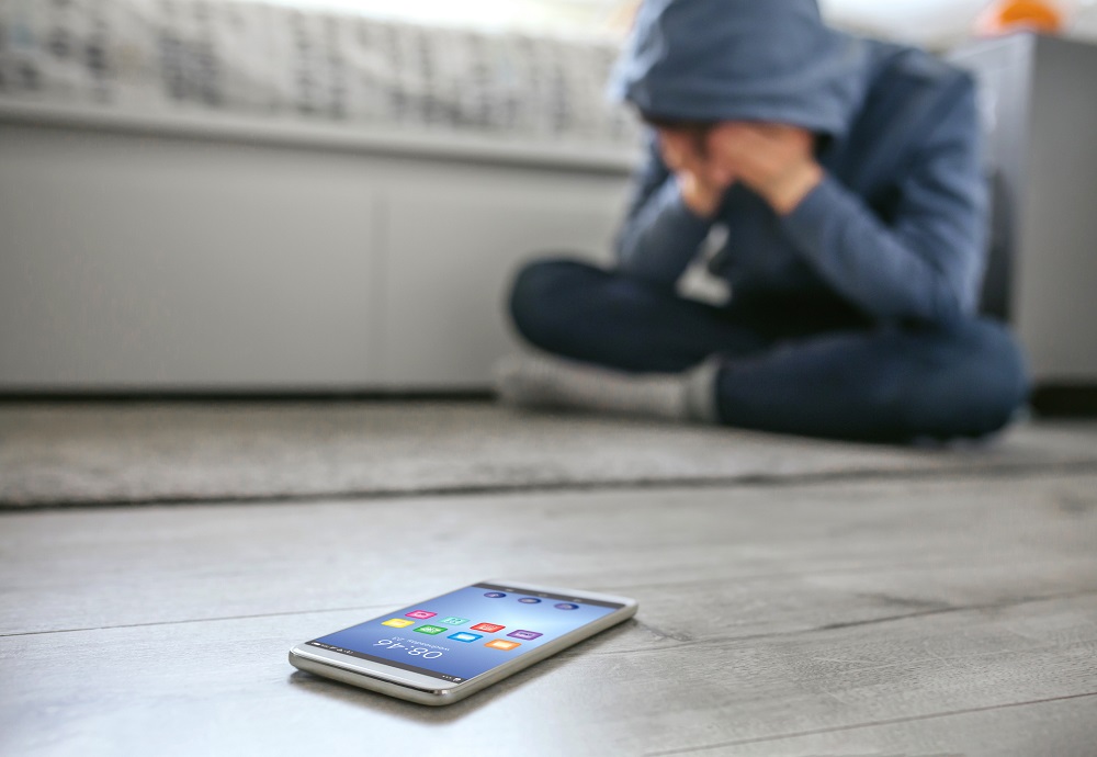 Teen boy sitting on his bedroom floor covering his face with his hands, his mobile phone on the floor in front of him