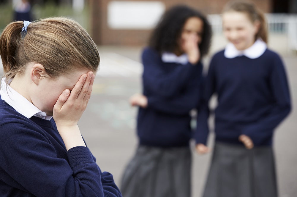 Girl in school uniform covering her face with her hands, with two other girls in the background talking about her behind their hands