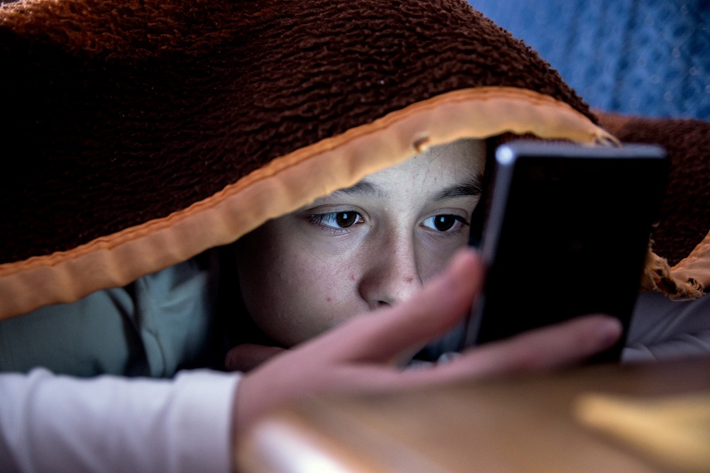 Young boy hiding under the bed covers looking at his mobile phone