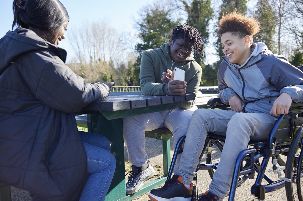 Happy teenage girl and two boys sitting outside laughing looking at a mobile phone. One boy is in a wheelchair.