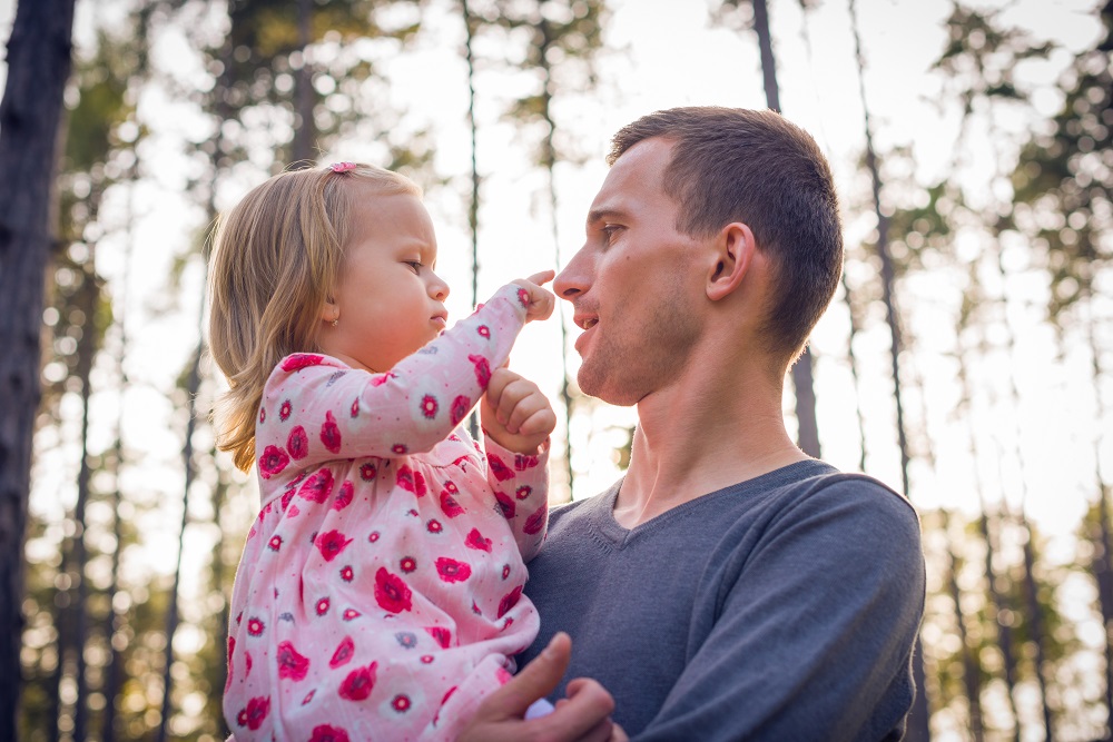 Dad holding toddler girl so they can chat face to face, outdoors in a wood