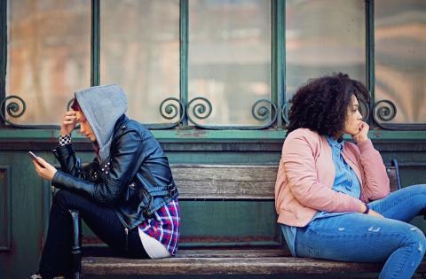 Two teenage girls sitting on a bench with their backs to each other, looking fed up. One girl is looking at her phone.